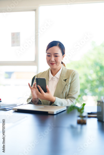 A smiling businesswoman in a light suit is seated at her desk, holding her phone in a bright office setting, looking engaged and connected