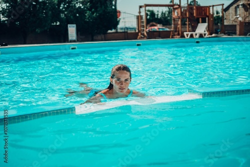 A woman is swimming in a pool. She is wearing a green top and is smiling. The pool is blue and has a few chairs around it.