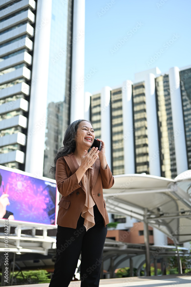 Fototapeta premium Cheerful mature businesswoman enjoying a phone conversation in front of skyscrapers