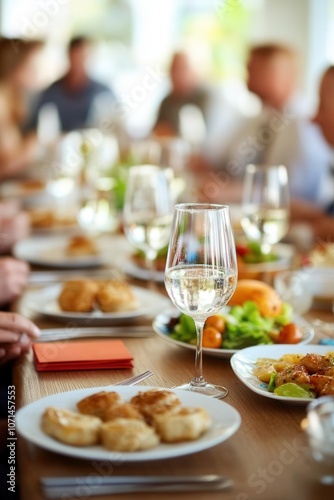 A glass of white wine sits on a table with food and other glasses as a group of people enjoy a meal together.