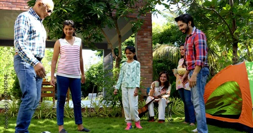 Indian Asian multigenerational family enjoying outdoor camping time as grandpa and son play football with granddaughters, while grandma and mother watch and cheer with smiles photo