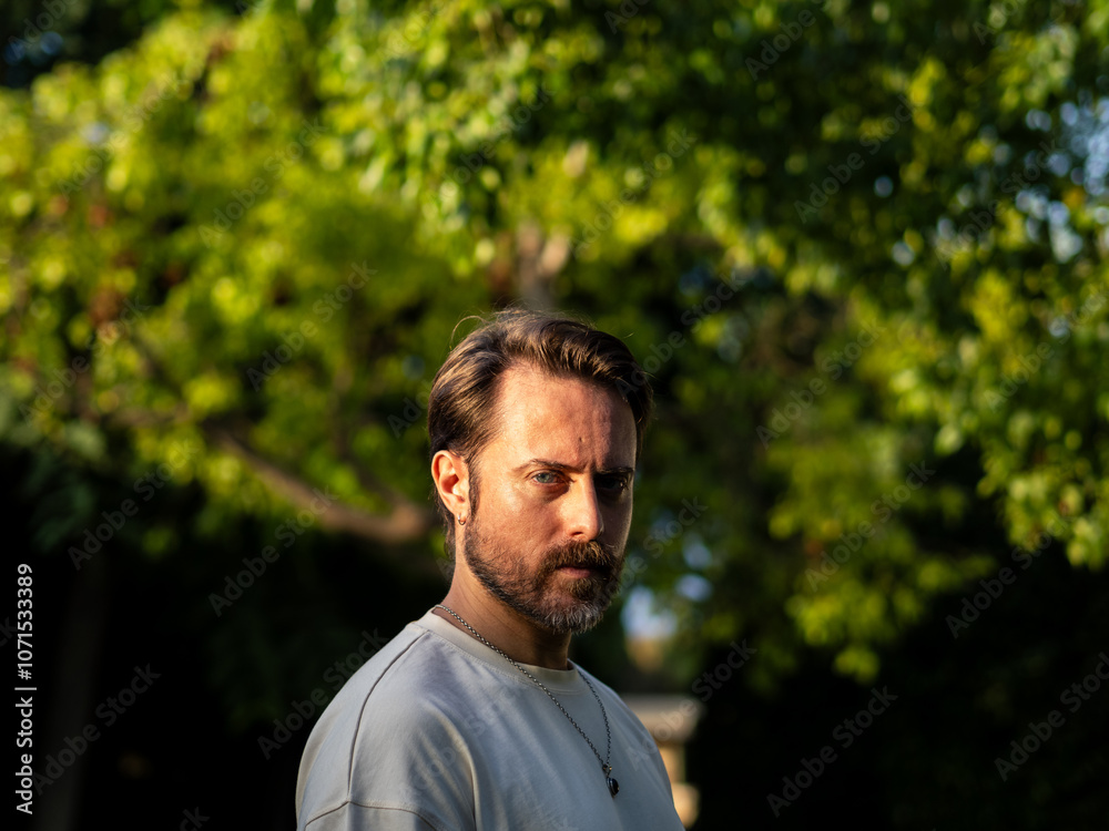 Intense portrait of a man with a beard looking at camera, surrounded by vibrant green trees in the late afternoon light, creating a serene and reflective atmosphere. Intense portrait of a man with a beard looking at camera, surrounded by vibrant green trees in the late afternoon light, creating a serene and reflective atmosphere.