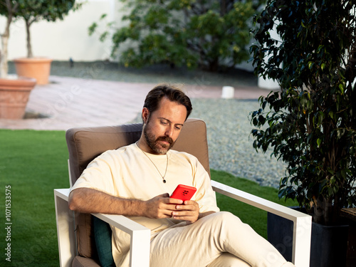 A man sits comfortably in an outdoor chair, focused on his smartphone. The setting features grass, potted plants, and a peaceful environment, perfect for relaxation in the sun. photo