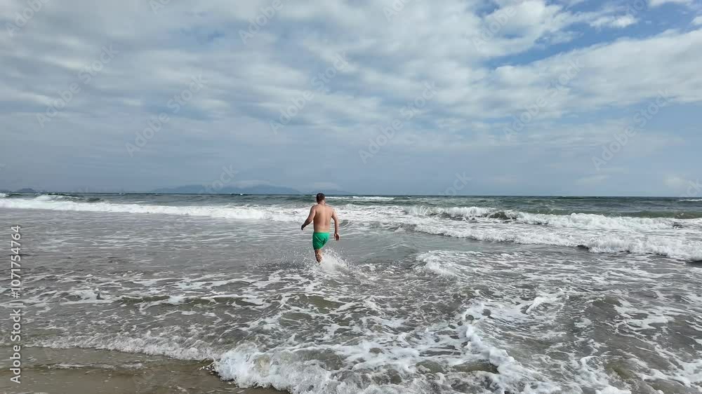 A Caucasian man in green shorts walks into the ocean on a cloudy summer day concepts include vacation and relaxation A Caucasian man in green shorts walks into the ocean on a cloudy summer day concepts include vacation and relaxation