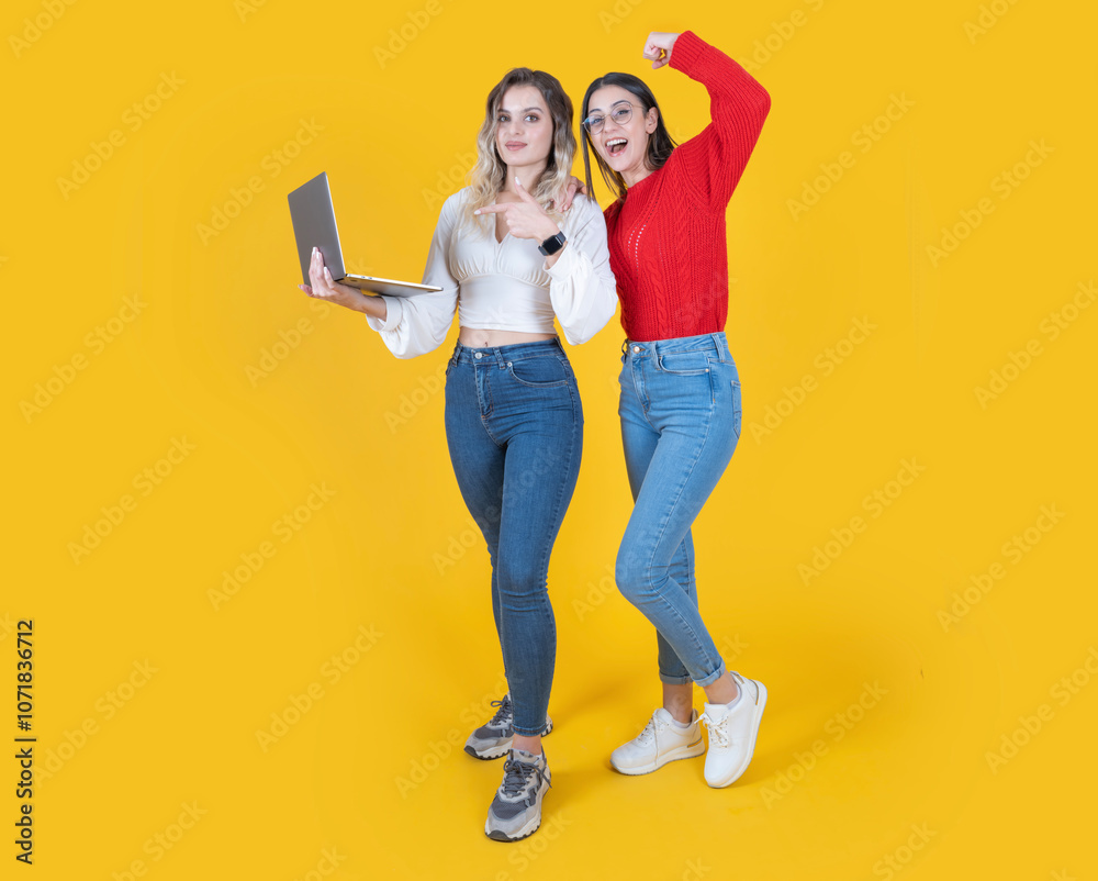 Fototapeta premium Using laptop, full body length two women standing isolated on yellow background holding using laptop pointing screen doing winner gesture wear casual clothes copy space.