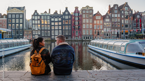 Photography A couple enjoys a serene moment by the canals in Amsterdam, surrounded by colorful, historic houses reflecting in the water, capturing the essence of autumn in this beautiful city