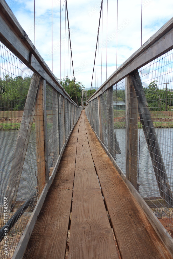 Obraz premium Rickety Swinging Wooden Bridge in Kauai, Hawaii (Hanepepe). Three wooden floor boards, windy. Cloudy skies.