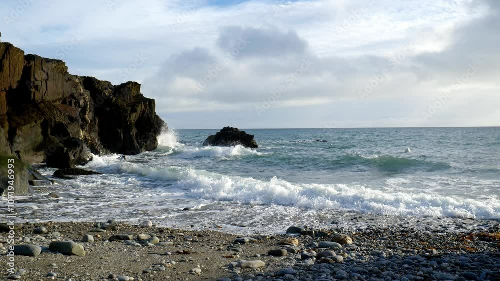 A large rock formation stands on the shore of the ocean. The water is choppy and waves are crashing against the rocks. There is a pebble beach in the foreground.