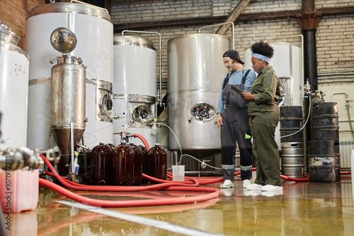 Wide angle shot of female African American process supervisor and male factory worker wearing denim overalls inspecting filling kegs with cider in workshop at plant photo