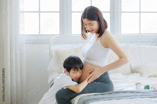 Pregnant mother and son are talking and spending time together in bed at home. Little child boy looking at her mother pregnant tummy. Pregnancy, family, preparation and expectation concepts. photo