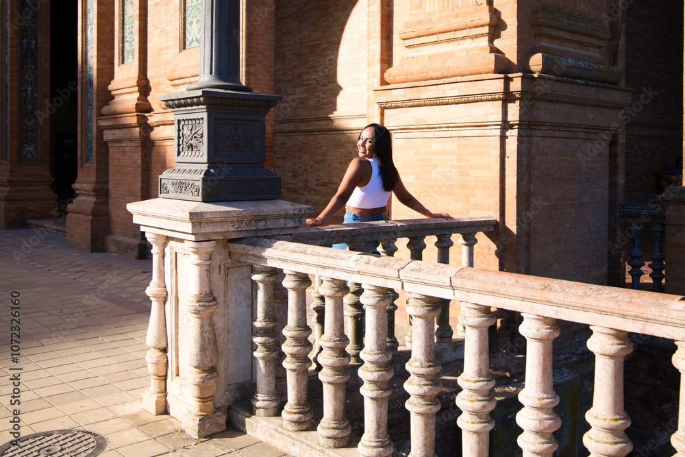 Fototapeta premium young and pretty brunette Moroccan woman dressed in white top and jeans is happy leaning on the railing in the square of Spain in Seville, she looks back and takes the photo.