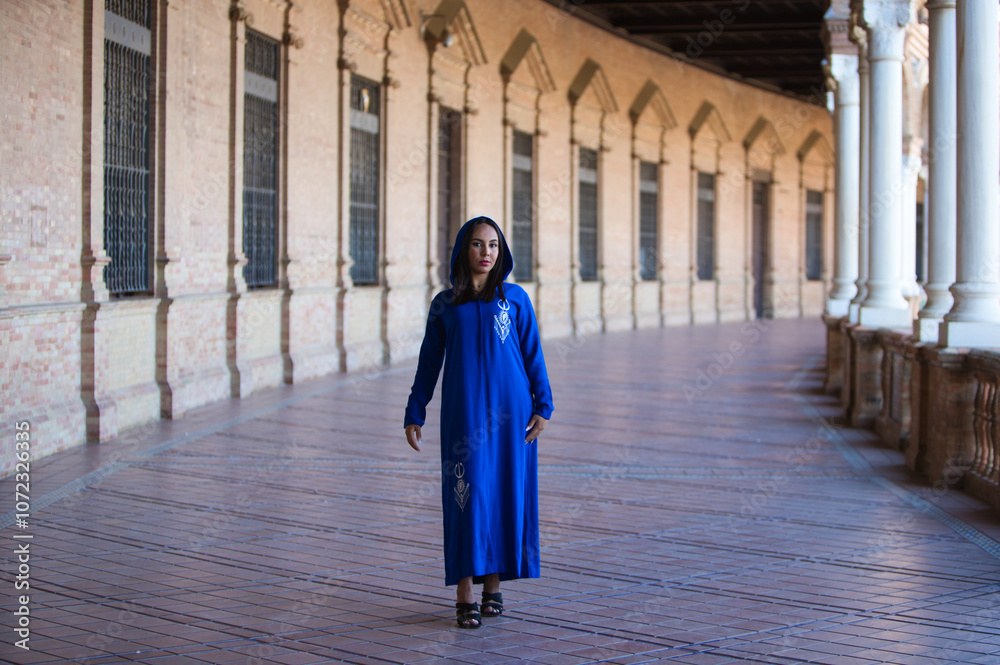 Naklejka premium young, beautiful, brunette moroccan woman, dressed in blue abaya, typical arabian costume, walks between arches and columns in the square of spain in Seville.The African woman is on holiday in Europe.