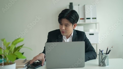 Asian businessman working at his office desk becomes increasingly frustrated while handling a phone call. Stress and pressure mount as he navigates multiple tasks on his laptop