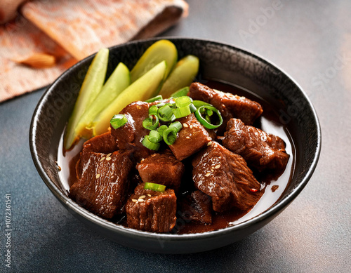 Asian-style braised beef cubes in dark soy sauce, garnished with green onions and sesame seeds, served with pickled cucumber slices in a black ceramic bowl