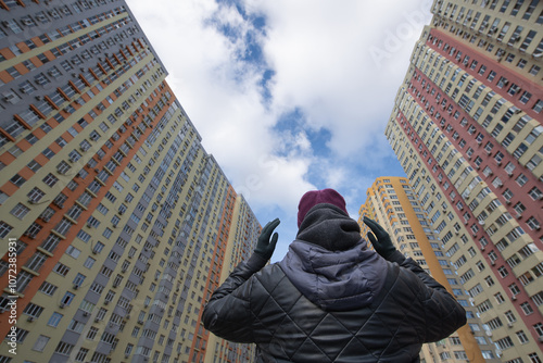 In an urban environment, an individual stands between tall residential buildings, looking contemplatively at the sky filled with clouds, showcasing city life’s contrast