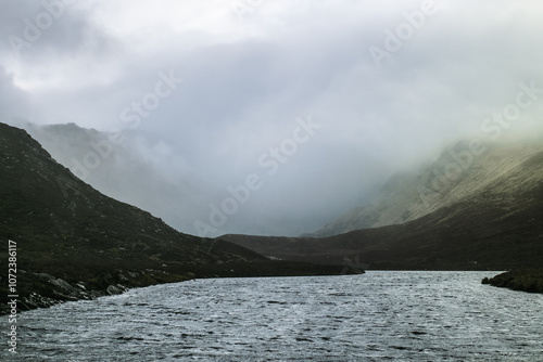 A serene river meanders through a mountainous area, shrouded in mist. The fog gently envelops the hills, creating a calm and peaceful atmosphere in the early morning.