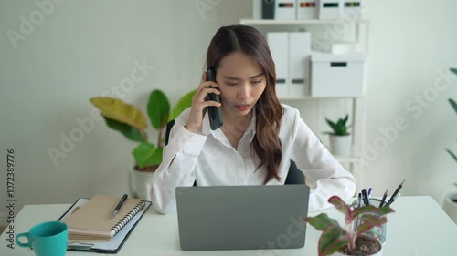 Young businesswoman is sitting at her desk, experiencing stress while working on her laptop. She is holding her phone and looking at the screen with a worried expression