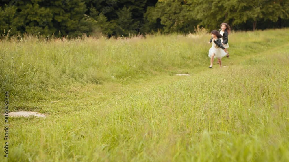 Two caucasian little girls in summer dress and black jacket playing together at summer garden. Pretty sisters in positive mood relaxing on fresh air. Two sisters run across the field Two caucasian little girls in summer dress and black jacket playing together at summer garden. Pretty sisters in positive mood relaxing on fresh air. Two sisters run across the field