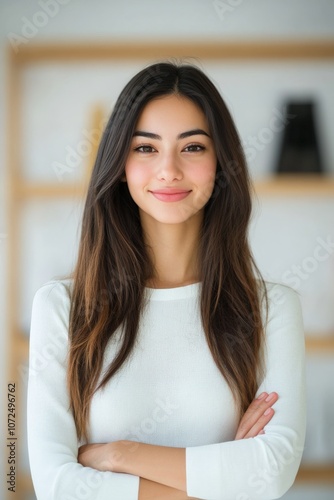 A young woman stands confidently, arms crossed, wearing a white top. Her long, straight hair and welcoming smile exude a sense of warmth and positivity.