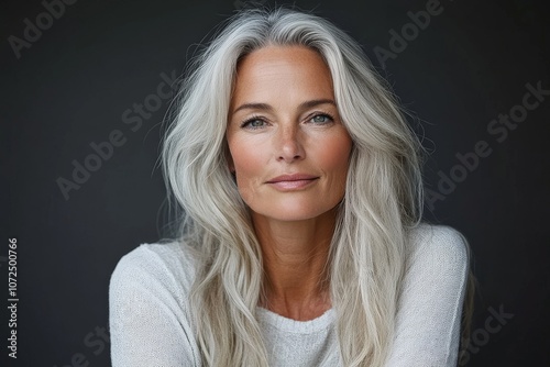 An elegant woman with long, flowing silver hair gazes calmly at the camera, conveying wisdom and elegance, set against a simple dark background indoors.