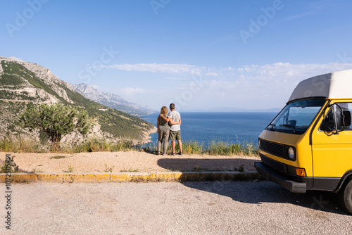 Couple enjoying the view of Croatian mountainous coast standing by their retro yellow camper van parked on a cliff, Croatia