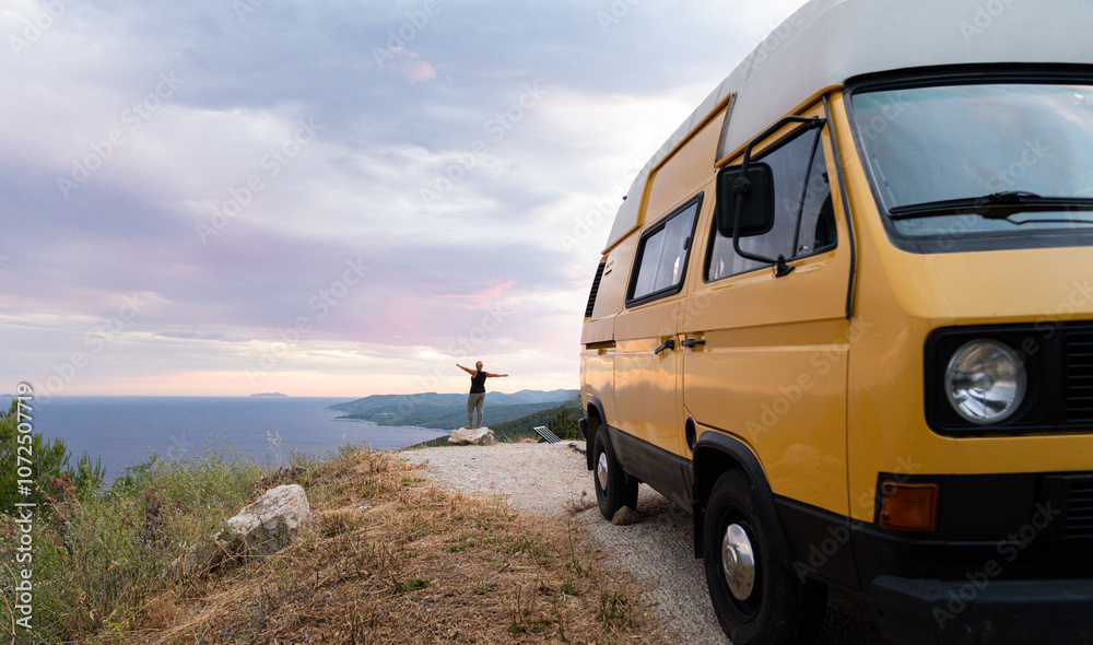 Yellow retro camper van parked on a dirt road above the sea on Korcula island, Croatia. Small female silhouette standing on the cliff with her hands up. Yellow retro camper van parked on a dirt road above the sea on Korcula island, Croatia. Small female silhouette standing on the cliff with her hands up.