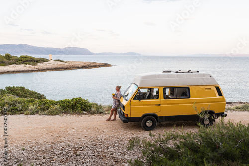 Young blonde woman enjoying the sea view in her yellow retro camper van parked on a dirt road above the sea on Korcula island, Croatia