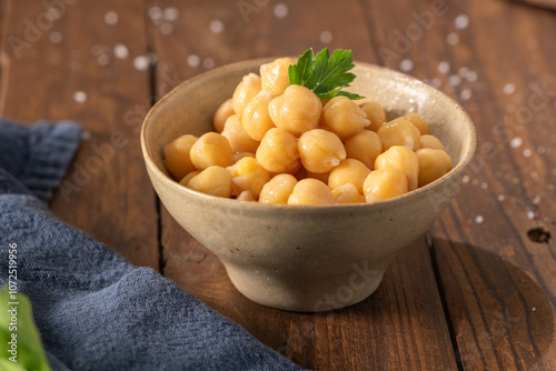 Cooked chickpeas in a ceramic bowl on a rustic wooden table decorated with a parsley leaf, ready to eat