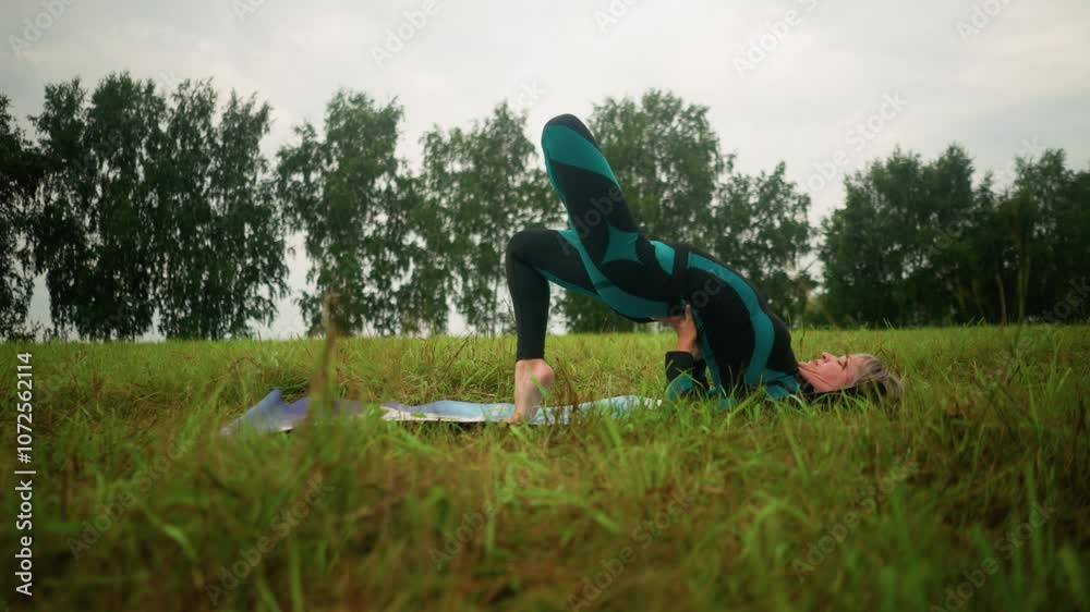 Middle-aged woman lying on a yoga mat practicing bridge pose with hands supporting her waist in a vast grassy field under cloudy skies, with trees lined in the distance Middle-aged woman lying on a yoga mat practicing bridge pose with hands supporting her waist in a vast grassy field under cloudy skies, with trees lined in the distance