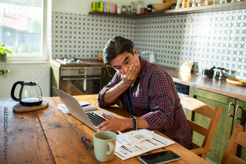 Stressed man looking at laptop with bills on tablet at kitchen photo