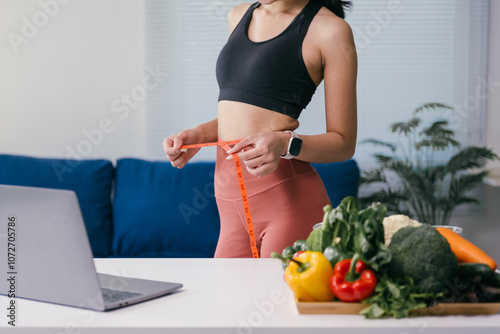 Young woman measuring her waist with a tape measure while standing near a table with fresh vegetables and a laptop, promoting a healthy lifestyle at home photo