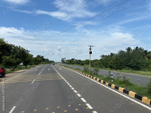 Scenic road in Kanyakumari, Tamil Nadu, India, bordered by lush greenery, under a vibrant blue sky. Captured on a sunny day, this picturesque highway leads through the tranquil landscape of southern I