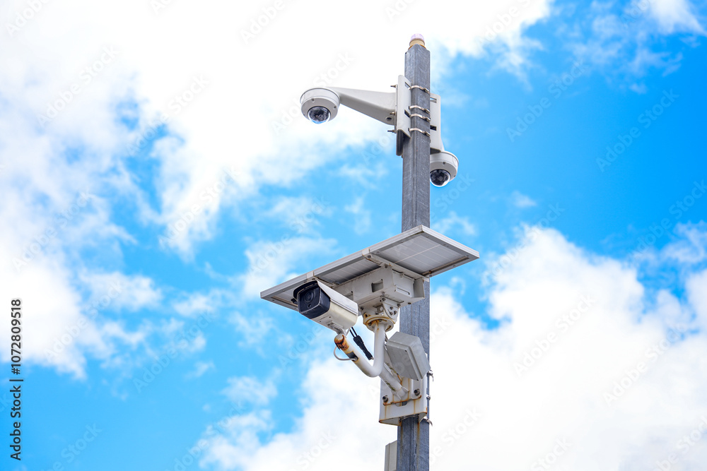 Modern Surveillance Camera on a Pole Against a Bright Blue Sky with Fluffy Clouds, Highlighting Security Technology and Outdoor Monitoring Solutions Modern Surveillance Camera on a Pole Against a Bright Blue Sky with Fluffy Clouds, Highlighting Security Technology and Outdoor Monitoring Solutions