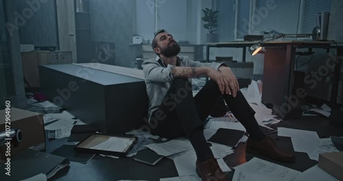 Dolly shot of depressed businessman sitting on floor after fit of rage over failed business deal or receiving bad news. Stressed office employee in destroyed office room. Financial crisis or firing.