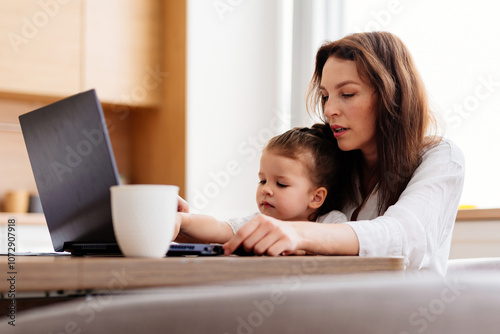 A woman works at a kitchen table with a laptop and a child