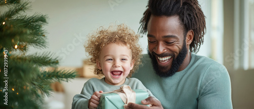 A joyful father and child share a special moment while unwrapping a gift near a decorated Christmas tree.