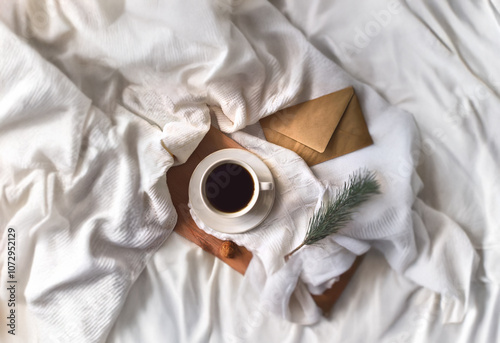 A cup of coffee, two envelopes, and a sprig of greenery rest on a wooden tray atop white bedding.