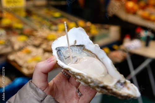 A huge oyster for sale at Tsukiji Outer Fish Market in Chuo, Tokyo, Japan - held up by a woman’s hand, with more fresh seafood for sale in the background photo