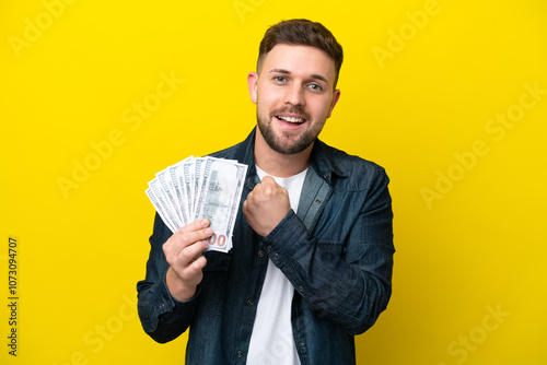 Young caucasian man taking a lot of money  isolated on yellow background celebrating a victory