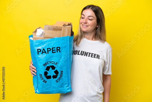 Young Rumanian woman holding a recycling bag full of paper to recycle isolated on yellow background with happy expression