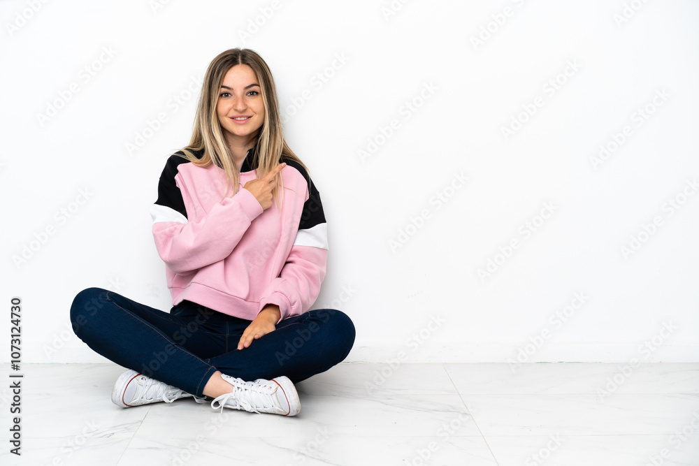 Young woman sitting on the floor at indoors pointing to the side to present a product