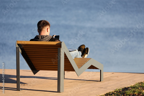 Fototapeta Naklejka Na Ścianę i Meble -  Man lying with smartphone in hands in deck chair on autumn beach. Holidays in cold weather