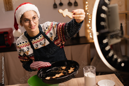 baker online courses. Smiling female blogger cooking gingerbread cookies and shooting video of herself using mobile phone on a tripod. Culinary training class photo