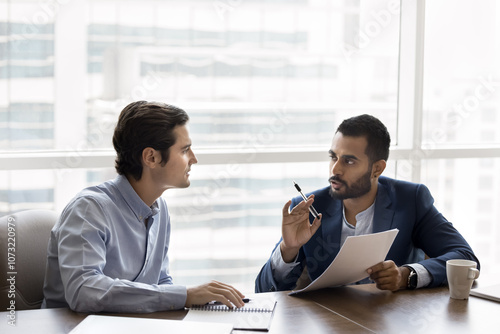 Two diverse serious young male business partners discussing contract terms, agreement text, marketing financial report, reviewing paper document at workplace table together photo