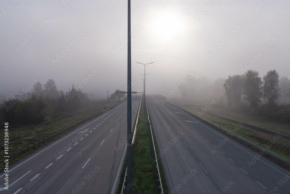 Dwujezdniowa, czteropasmowa autostrada. Nad jezdnią unosi się mgła ograniczająca widoczność. Jest ona od przodu oświetlana promieniami słońca.