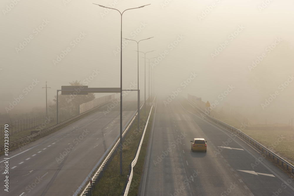 Dwujezdniowa, czteropasmowa autostrada. Nad jezdnią unosi się mgła ograniczająca widoczność. Jest ona od przodu oświetlana promieniami słońca.