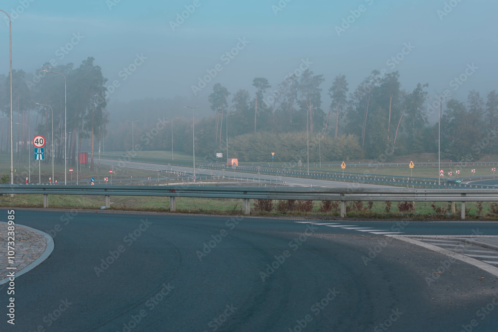 Węzeł drogowy przy autostradzie. Widać zjazdy i rozjazdy. Nad węzłem drogowy jak i nad pobliskim lasem unosi się mgła.