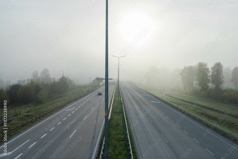 Dwujezdniowa, czteropasmowa autostrada. Nad jezdnią unosi się mgła ograniczająca widoczność. Jest ona od przodu oświetlana promieniami słońca.