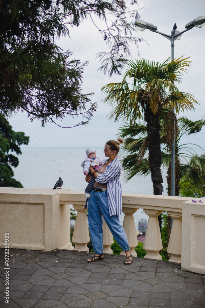 Mom and baby - little daughter in their arms in the park against the background of the sea and palm trees Mom and baby - little daughter in their arms in the park against the background of the sea and palm trees