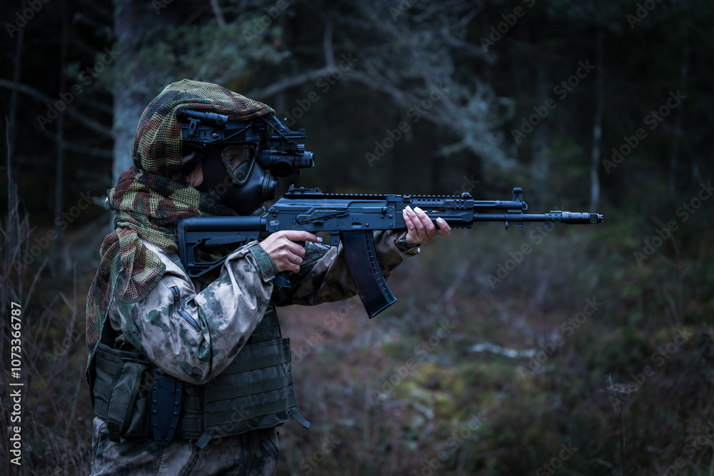 Fototapeta premium A woman soldier in camouflage, wearing gas mask and a helmet with a night vision device, assault rifle in her hands in a dark forest.